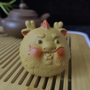 May include: A ceramic figurine of a round, yellow creature with red accents on its cheeks and a small orange crest. The figurine has small, dark eyes and a slightly grumpy expression. It is sitting on a bamboo tray, with a brown teapot in the background.