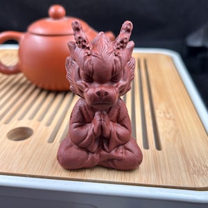 May include: A reddish-brown dragon figurine in a seated position with hands clasped in prayer. The dragon has detailed features, including horns and scales. A matching teapot is in the background, resting on a wooden tray.