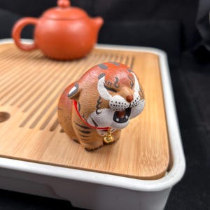 May include: A ceramic tiger figurine with orange and brown stripes, a red collar, and a small gold bell. The tiger is on a wooden tray, with a terracotta teapot in the background. The tiger has its mouth open.