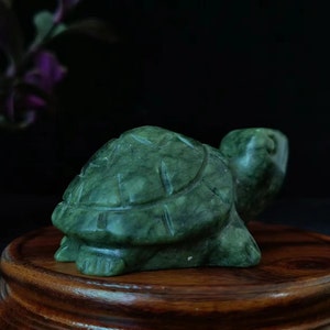 Può includere: Statuetta di tartaruga in giada scolpita, nei toni del verde. La tartaruga presenta linee incise sul guscio, zampe e testa visibili. La statuetta è esposta su un supporto rotondo in legno, su sfondo scuro.