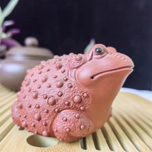 Puede incluir: Figura de rana de cer&aacute;mica de color marr&oacute;n rojizo con una superficie texturizada y con bultos. La rana tiene una cara detallada con un ojo peque&ntilde;o y oscuro y est&aacute; sentada sobre una superficie de madera. Una tetera es visible en el fondo.