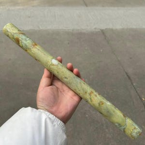 May include: A cylindrical, light green stone rod with brown and white marbling. The stone is held in a hand, showcasing its smooth, polished surface. The rod appears to be approximately 10 inches long.
