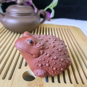 Puede incluir: Una figura de rana de cer&aacute;mica de color marr&oacute;n rojizo con una superficie texturizada y con bultos. La rana tiene una cara detallada con un ojo oscuro y est&aacute; colocada sobre una bandeja de bamb&uacute;. Una tetera marr&oacute;n est&aacute; en el fondo.