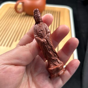 Puede incluir: Pequeña estatua de color marrón rojizo de una figura vestida con una túnica y un tocado adornado, sostenida en una mano. La figura está de pie sobre una pequeña base rocosa. Una tetera y una bandeja son visibles en el fondo.
