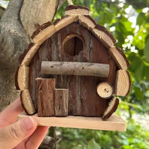 Puede incluir: Un nido de pájaros rústico de madera con acabado de corteza natural. Presenta un orificio de entrada redondo, una pequeña percha y una puerta en miniatura. El tejado y los bordes están adornados con piezas de madera clara. Diseñado para colgar en exteriores.