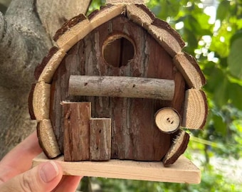 Puede incluir: Un nido de pájaros rústico de madera con acabado de corteza natural. Presenta un orificio de entrada redondo, una pequeña percha y una puerta en miniatura. El tejado y los bordes están adornados con piezas de madera clara. Diseñado para colgar en exteriores.