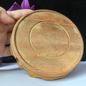 Puede incluir: Un posavasos redondo de madera clara. Presenta una hendidura circular central y un borde elevado. La veta de la madera es visible, aportando textura. El posavasos está diseñado para sostener una taza o un vaso. Diámetro aproximado 10 cm.