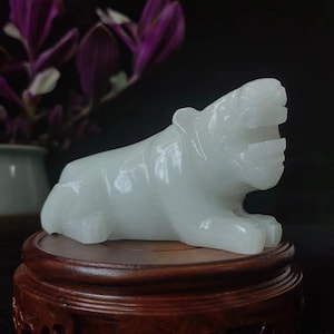 May include: A white jade tiger sculpture, lying down with its mouth open, is displayed on a round, dark wooden stand. The tiger is carved with detailed features and sits against a dark background, showcasing the sculpture's smooth texture.