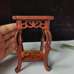 May include: A miniature, ornate wooden table with a reddish-brown finish. The table features intricate cut-out designs on the sides and a lower shelf. The legs have a curved, elegant shape, and the overall style is traditional.