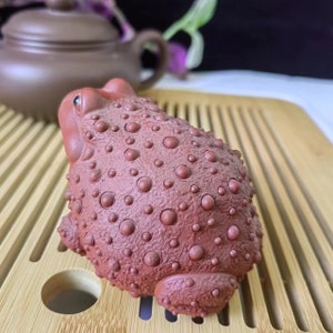 Puede incluir: Figura de rana de cer&aacute;mica marr&oacute;n rojizo con una superficie texturizada y con bultos. La rana est&aacute; sobre una bandeja de madera clara, con una tetera marr&oacute;n al fondo. La figura tiene una apariencia detallada y realista.