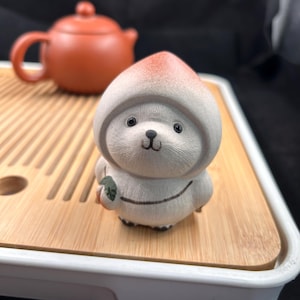 May include: A ceramic figurine of a bear-like creature wearing a hooded outfit with a peach-colored top. The figurine is holding a small green object. A small, orange teapot is in the background, resting on a bamboo tray.