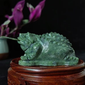 May include: A detailed, green jade frog sculpture with a long tongue extended, resting on a wooden stand. The frog has textured skin and is set against a dark background with purple flowers. The sculpture is a decorative item.