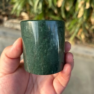 May include: A cylindrical, dark green stone object with a polished surface. The stone has natural variations in color and texture, and is held in a hand. The object appears to be a small cup or container.