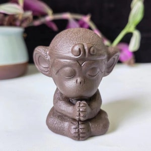 Peut inclure: Figurine de singe en céramique marron, assis, les mains jointes en prière. Le singe a les yeux fermés et une surface texturée détaillée. L'objet est décoratif, possiblement pour la décoration intérieure ou les pratiques spirituelles.