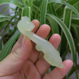 May include: A pale green jade carving of a stylized animal, possibly a dragon or mythical creature. The carving is smooth and elongated, with detailed features. The object is held in a hand against a background of green foliage.