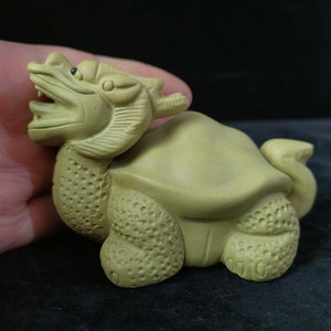 May include: A carved, light green figurine of a mythical creature, a hybrid of a turtle and a dragon. The figurine has a detailed, textured surface, with the dragon's head featuring an open mouth and detailed eyes. The turtle-dragon is posed on a dark surface.