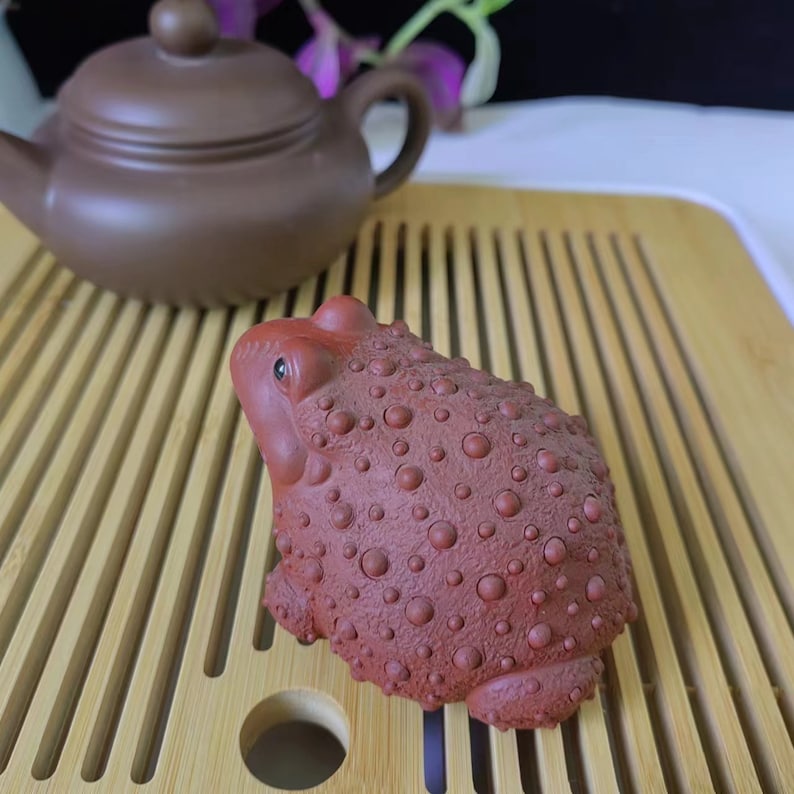 Puede incluir: Figura de rana de cer&aacute;mica de color marr&oacute;n rojizo con una superficie texturizada y con bultos. Una tetera marr&oacute;n se encuentra en el fondo sobre una bandeja de bamb&uacute;. La rana es un objeto decorativo, posiblemente para una ceremonia del t&eacute;.