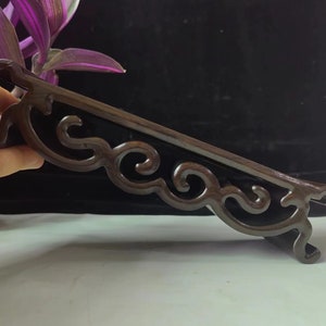 May include: A dark brown wooden display stand featuring a smooth, polished finish and an intricate, openwork carved design along the side. The stand has a flat top surface and curved, scroll-like legs that provide a stable base for objects.