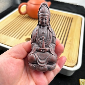 May include: A reddish-brown statue of a seated Buddha figure with hands in a prayer position. The statue has detailed facial features and flowing robes. The figure is approximately 3 inches tall.