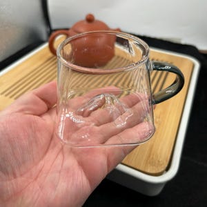 May include: A clear glass teacup with a mountain-shaped base and a dark green handle. The teacup is held in a person's hand, with a brown teapot and bamboo tray in the background. The teacup is designed for tea.