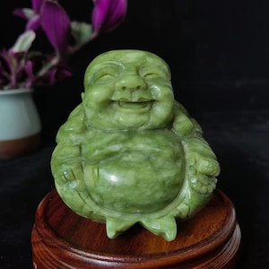 Può includere: Una statua di Buddha sorridente in giada verde. La figura è scolpita con dettagli precisi e poggia su un supporto rotondo in legno. La giada presenta variazioni naturali di colore ed è un simbolo di pace e prosperità.