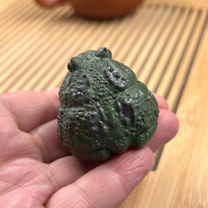 May include: A small, dark green toad figurine with a textured, bumpy surface and darker blackish-green mottled patches across its back. The toad is depicted in a seated, hunched position with its head slightly lowered and eyes visible at the top.