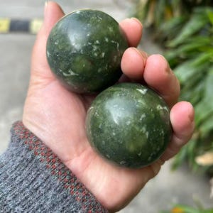 Peut inclure: Deux pierres sphériques lisses avec une finition polie. Les pierres sont d'un vert forêt profond, présentant des inclusions minérales naturelles blanches et vert clair ainsi que des veines sur leurs surfaces arrondies, créant un aspect marbré.