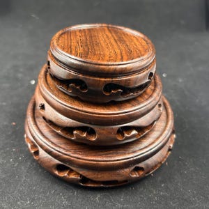 Könnte beinhalten: Ein Stapel von drei dunkelbraunen Holz-Ausstellungsständern. Jeder Ständer hat eine runde Oberseite und geschnitzte wellenartige Designs an den Seiten. Die Holzmaserung ist auf den Oberflächen sichtbar. Ideal für Dekorationsartikel.