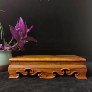 May include: A rectangular wooden display stand with a rich, reddish-brown finish. The stand features a decorative, wave-like design along the bottom edge. The stand is likely for displaying small objects. A purple plant is in the background.