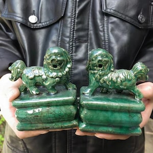 Może przedstawiać: Dwie pasujące zielone ceramiczne posągi Foo Dog. Każdy pies siedzi na prostokątnej podstawie z efektem spękanego szkliwa. Psy mają szczegółowe cechy, w tym uśmiechniętą minę i płynącą grzywę.