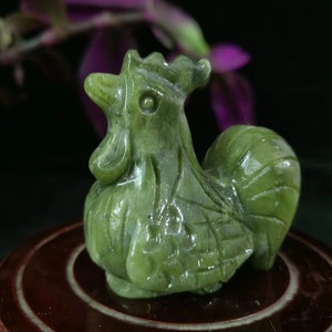 Può includere: Una statuetta di gallo in giada scolpita, nei toni del verde. La scultura presenta dettagli, tra cui cresta, bargigli e penne della coda. Il gallo è posizionato su una base circolare marrone scuro, con uno sfondo sfocato di fiori viola.