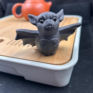 May include: A small, dark gray ceramic bat figurine with pointed ears, wide outstretched wings, and two visible fangs. The bat has a textured body with a carved heart-shaped design on its chest and glossy black eyes, positioned in a standing pose.