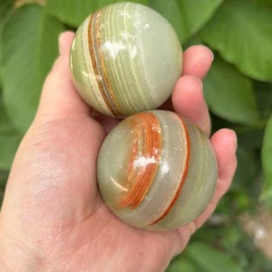 May include: Two polished onyx spheres, each displaying bands of green, brown, and orange hues. The spheres are held in a hand, with a backdrop of green foliage. The smooth, rounded stones are a natural decorative item.