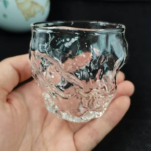 Peut inclure: Un gobelet en verre transparent avec un design texturé et organique. Le verre présente une surface rugueuse et inégale, créant un effet visuel distinctif. Le gobelet est tenu dans une main, mettant en valeur sa taille et sa forme. Le design suggère un produit artisanal.