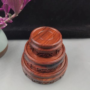 Puede incluir: Una pila de tres soportes de exhibición redondos de madera. Los soportes son de un rico color marrón rojizo con intrincados diseños tallados alrededor de los bordes. El soporte superior es el más pequeño y el inferior es el más grande.