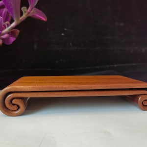 May include: A small, rectangular wooden display stand with a rich, reddish-brown finish. The stand features a curved, scroll-like design on each end, adding a touch of elegance. The stand is ideal for displaying small items.
