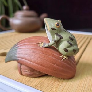 Puede incluir: Una figura de cerámica que representa una rana de color verde claro con manchas de color marrón oscuro posada sobre un objeto curvo, texturizado y de color marrón rojizo que recuerda a una hoja o una concha. La rana tiene detalles dorados en los dedos.