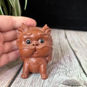 May include: A small, brown figurine of a mythical creature with large eyes, horns, and decorative swirls. The figurine is standing on a wooden surface. The creature has a surprised expression.