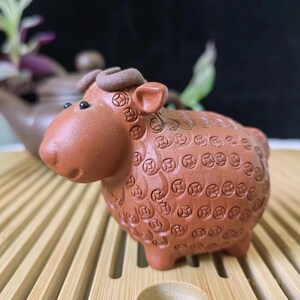 May include: A terracotta sheep figurine with detailed coin-like patterns on its body. The sheep has small, curved horns and black eyes. The figurine is a decorative item, likely for home decor or a collection.