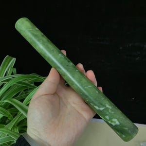 May include: A green jade roller, held in a hand, against a dark background. The cylindrical roller is a solid green colour with white marbling. The roller is approximately 20 cm long.