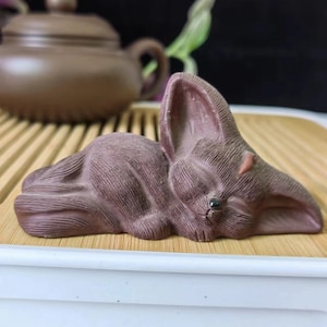 以下が含まれることがあります： 長い耳を持つ、眠っているウサギの茶色のセラミック彫刻。 ウサギは竹のトレイの上に横たわっており、背景には茶色のティーポットがあります。 彫刻は質感のある表面と小さな黒い目を持っています。