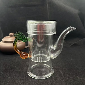 May include: Clear glass teapot with a decorative handle in shades of green and orange. The teapot has a spout and a built-in infuser. A brown teapot is visible in the background.