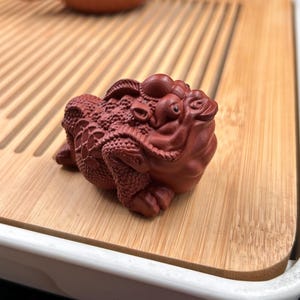 May include: A reddish-brown clay figurine of a three-legged toad with a textured, bumpy back and detailed scales. The creature has a wide, expressive face with prominent eyes and a small mouth, crafted with a matte finish and intricate carvings.