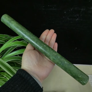 May include: A smooth, cylindrical jade roller in shades of green is held in a hand. The jade roller is approximately 20 cm long. The background is dark, with green and white plants visible on the left side.