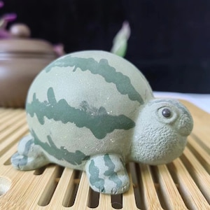 May include: A ceramic turtle figurine with a watermelon design. The turtle is light green with dark green stripes, and has a detailed face and feet. The figurine is sitting on a bamboo tray.