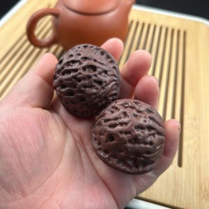 May include: Two brown, walnut-shaped objects with textured surfaces are held in a hand. A brown teapot and a wooden tea tray are in the background. The objects appear to be decorative or possibly used in a tea ceremony.