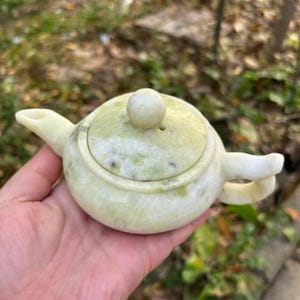 Könnte beinhalten: Kleine, hellgrüne Teekanne aus Stein mit rundem Körper, Ausguss und Henkel. Der Deckel hat einen kleinen, runden Knopf. Die Teekanne ist hellgrün mit dunkleren grünen und braunen Adern. Ein dekorativer Artikel.
