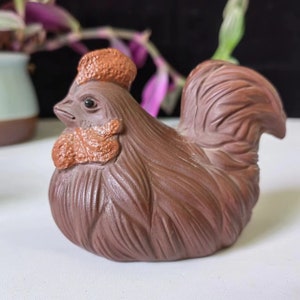 Può includere: Statuetta di gallo in ceramica marrone con dettagliate texture di piume e cresta. Il gallo ha un corpo arrotondato e una testa dettagliata, con cresta e bargigli rosso-brunastri. Oggetto decorativo.