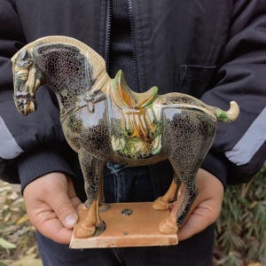 May include: A ceramic horse figurine with a saddle. The horse is mainly black with a crackle glaze, and the saddle has green and tan accents. The figurine is mounted on a rectangular base.