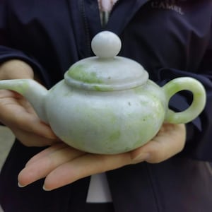 Puede incluir: Una pequeña tetera verde pálido con tapa a juego y asa redondeada. La tetera está hecha de un material de piedra y tiene un pomo blanco en la parte superior de la tapa. La tetera está sostenida en las manos de alguien.